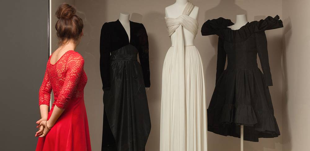 Blick in die Modegalerie – Person in rotem Kleid betrachtet drei Schaufensterpuppen in schwarzem und cremefarbenem Kleid. Fotografie.