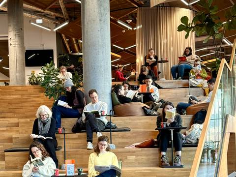 Foto: Pausify – Personen auf Holztribüne in modernem Raum, lesen konzentriert Bücher. Pflanzen und Glaswand im Hintergrund. Entspannte Atmosphäre mit Sitzkissen.
