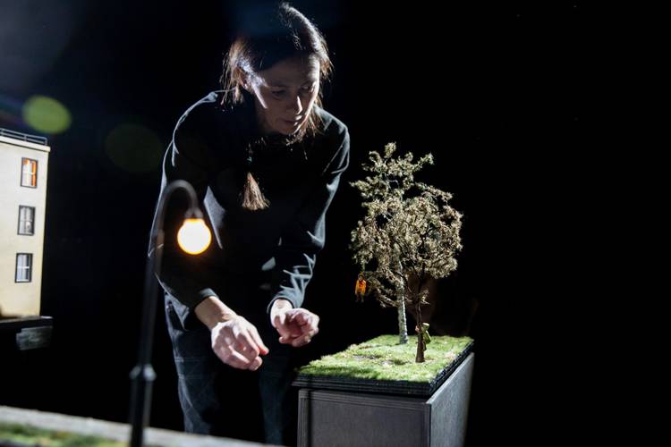 Transport: Departure – Person mit langen Haaren beugt sich über Miniaturlandschaft mit Baum und Figur. Im Hintergrund Gebäude und Laterne. Dunkler Hintergrund.