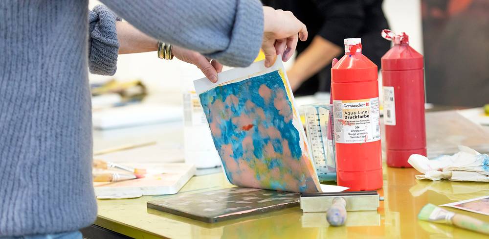 Person hält ein Blatt mit buntem Druck. Zwei rote Flaschen mit Aufschrift "Gerstaecker Aqua-Linol Druckfarbe". Malutensilien auf gelbem Tisch.