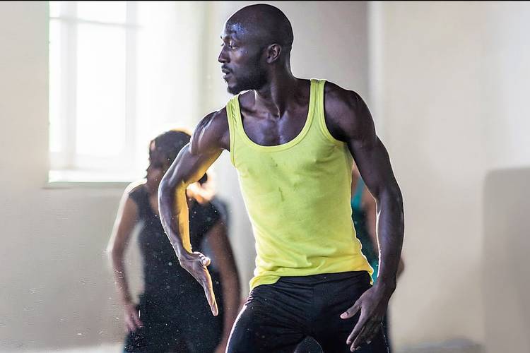 African traditional and afro-somatic dance – Person in gelbem Tanktop tanzt dynamisch, im Hintergrund verschwommen eine weitere Person. Fenster mit hellem Licht auf der linken Seite.