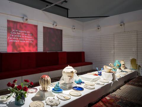  – Rote Sitzbänke an der Wand, darüber rotes Licht mit Text. Langer Tisch mit Kunstobjekten wie Vasen und Tellern, rote Rosen im Vordergrund.
