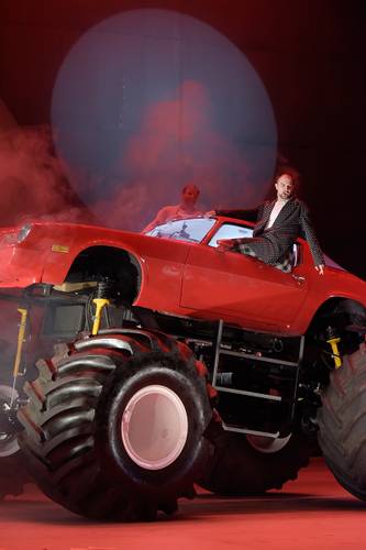 Die Entführung aus dem Serail – Roter Monstertruck, darauf sitzt eine männlich gelesene Person im Anzug; weitere Person am Steuer im Hintergrund; umgeben von rotem Scheinwerferlicht und Rauch.