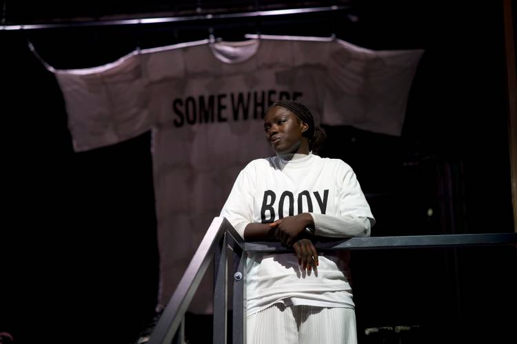 Foto: Lee Everett Thieler – Person mit T-Shirt "BODY" an Metallgeländer, im Hintergrund großes Shirt mit "SOMEWHERE" hängend.