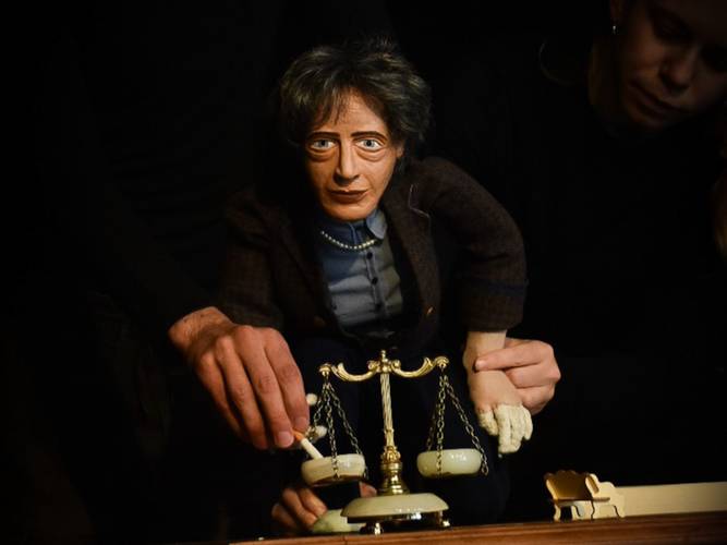 Wohl dem, der keine Heimat hat – Puppe mit realistischen Zügen, stehend vor einer Waage aus Spielzeug. Zwei Hände führen die Puppe. Person im Hintergrund in Schwarz, teilweise sichtbar.