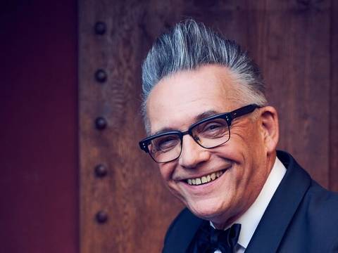 – Männlich wirkende Person mit grau meliertem Haar und Brille lächelt in die Kamera. Person trägt Anzug und Fliege. Hintergrund aus Holz.