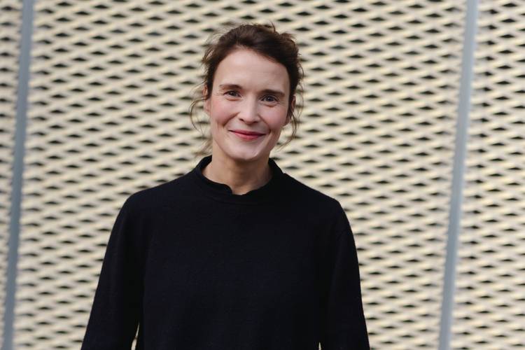 "ZEIT-Recherchen" mit Caterina Lobenstein – Eine Person mit dunklem Pullover lächelt vor einem gelochten Hintergrund.