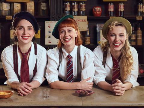 The Airlettes: Live – Drei lachende Personen in Hemden, Krawatten und Trägern hinter Tresen. Zwei tragen Baskenmützen, eine trägt einen hellgrünen Hut. Tisch hat Schale und Gläser.