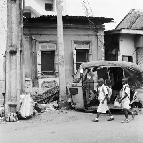 Akinbode Akinbiyi, Lagos Island, Lagos, 2016, Aus der Serie: „Lagos: All Roads“, seit den 1980er Jahren – Schwarz-weiß-Fotografie von zwei Schulkindern, die von rechts in das Bild laufen. Im Hintergrund sind das Gerüst eines kleinen Fahrzeugs und eine Hausfassade zu sehen.