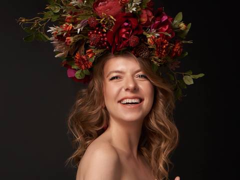  – Person mit langem, gewelltem Haar. Großer Blumenkranz aus roten und orangefarbenen Blumen mit grünem Laub auf dem Kopf. Lächelnder Ausdruck.
