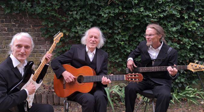 Drei Personen in schwarzen Anzügen mit Gitarren vor einer mit Efeu bewachsenen Mauer. Zwei sitzen, eine steht. Gitarren: Yamaha und klassische Gitarre.