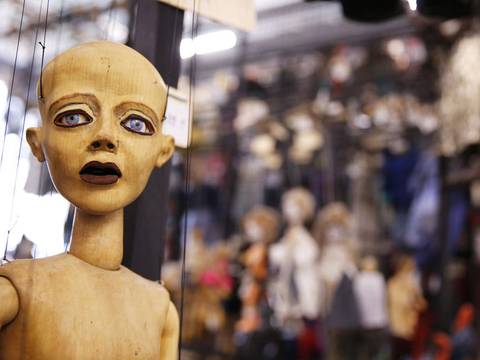  – Holzmarionette mit ausdrucksvollem Gesicht, groß im Vordergrund. Hintergrund unscharf mit vielen hängenden Puppen.