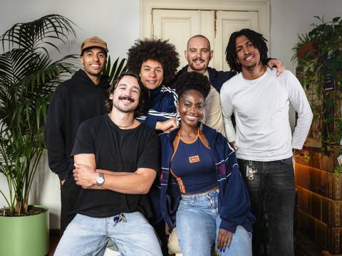 SACODE. Berlin-based collective rooted in afro:diasporic electronic music. – Sechs junge Personen lächeln vor einer Tür. Links steht eine grüne Pflanze.
