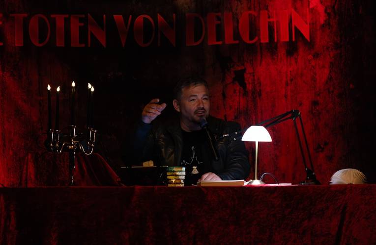 Männlich gelesene Person an Tisch mit Mikrofon und Lampe. Hintergrund: Schrift "TOTEN VON DELCHIN" auf rotem Vorhang. Links ein Kerzenständer.