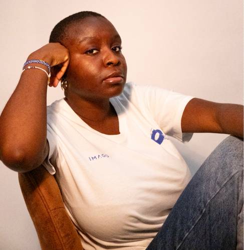 Person in weißem T-Shirt mit Aufschrift "IMAGO", sitzt, stützt den Kopf auf der Hand, schaut ernst, trägt Armbänder und Ohrringe.