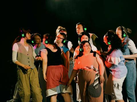 FRISCH gefragt!, junges RZt, RambaZamba Theater Berlin – Ein Bühnenfoto mit etwa Zehn bunt angezogenen Personen, die in einem dichten Pulk zusammen stehen. Alle tragen grüne Kopfhorer, wie bei einer Silent-Disko. Die Personen wirken Interresiert und musstern sich gegenseitig. Der Hintergrund ist schwarz.