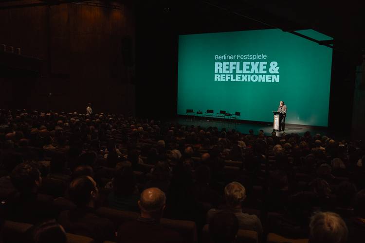 Publikum in einem Theater, Person an einem Rednerpult, Bildschirmtext: "Berliner Festspiele REFLEXE & REFLEXIONEN".