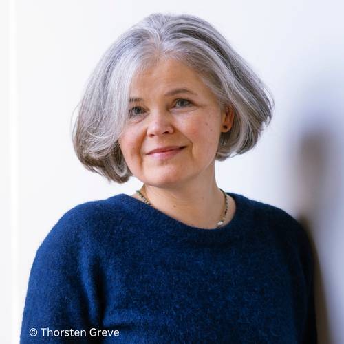 Julia Franck – Ältere Person mit grauen Haaren, blaue Strickpullover. Hintergrund weiß. Text: © Thorsten Greve.