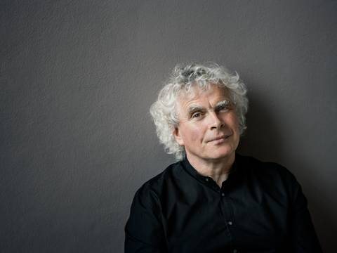 Sir Simon Rattle – Ein Mann mit weißen Locken und einem schwarzen Hemd steht vor einer neutralen Wand. Sein Blick in die Kamera ist entspannt.