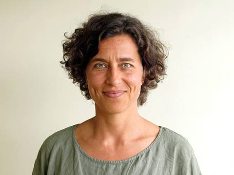 Theresa Adam – Lächelndes Gesicht einer Frau mit lockigem Haar, die ein graues Oberteil trägt, vor hellem Hintergrund.