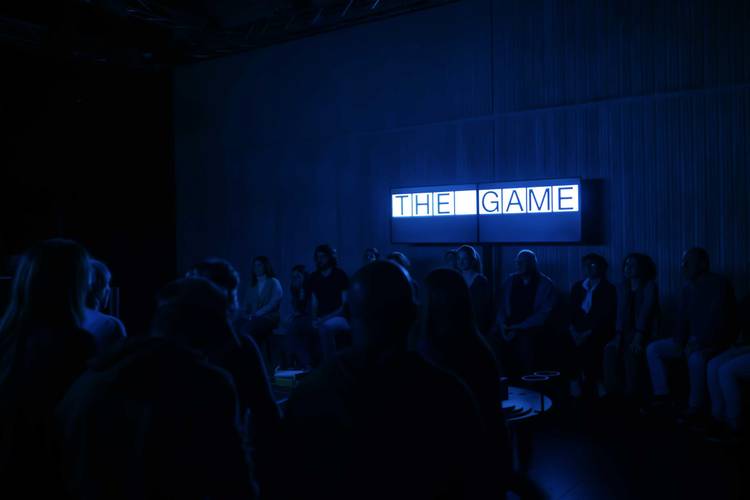 The Game – Menschen sitzen im Dunkeln vor einem leuchtenden Schild mit der Aufschrift "THE GAME".