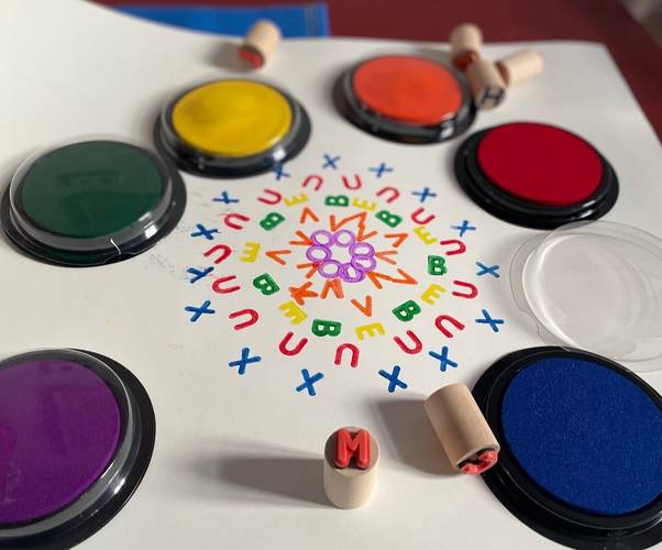 Mit Buchstabenstempeln und Farben entstehen bunte Muster. – Ein weißes Blatt mit farbig gestempelten Buchstaben sowie Stempelutensilien