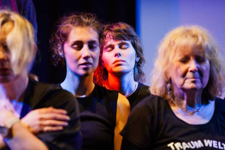 Einblicke: Workshop – Mehrere Personen mit geschlossenen Augen in dunkler Kleidung, bunte Beleuchtung. Vorderste Person trägt T-Shirt mit Schriftzug "TRAUM WE".