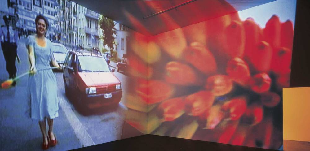 Pipilotti Rist, Ever is over All, 1997, Zweikanal-Video (color, sound), Videoinstallationmit zwei sich überlappenden Projektionen – Videoprojektion von Person mit Blume auf Straße, neben rotem Auto. Rechts große, unscharfe Darstellung roter Tulpen. Urbaner Hintergrund, Farbe.