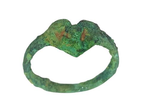 Kupferner Ring mit einem Herz und Initialen aus dem Zwangsarbeitslager Berlin-Köpenick – Ein schmaler, grünlich oxidierter Ring mit unregelmäßiger Oberfläche. Die Ringschiene ist dünn und wirkt stellenweise uneben. Auf der Vorderseite befindet sich eine herzförmige Platte mit zwei eingravierten, senkrechten Linien. Die Patina deutet auf ein altes oder antikes Schmuckstück hin.