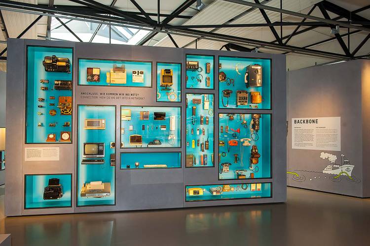 Hier wird die große Vielfalt historischer und aktueller Endgeräte gezeigt: vom Zeigertelegraf aus dem 19. Jahrhundert bis zu einer heutigen Smartwatch. – Eine große Vitrinenwand. Viele rechteckige, kleine und große Fenster geben den Blick frei auf Endgeräte aus unterschiedlichen Epochen. Die Rückwand der Vitrine ist strahlend blau.