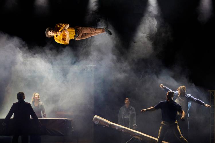 Kintsugi – Eine fliegende, männlich gelesene Person über vier Personen in Bühnenbeleuchtung und Rauch.
