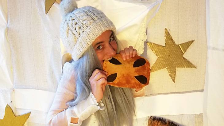 Knusper, Knusper - Hänsel und Gretel treffen die Weihnachtshexe Befana – Person mit weißer Strickmütze und langem grauem Haar hält Kissen mit schwarzen Flecken, umgeben von goldenen Sternen auf weißem Hintergrund.