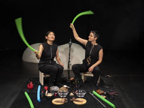 unsichtbar – Zwei Personen mit grünen Röhren auf Hockern. Verschiedene Gegenstände auf dem Boden: Schüssel, Bürste, rotes Horn, Trommel, blaue und grüne Röhren.