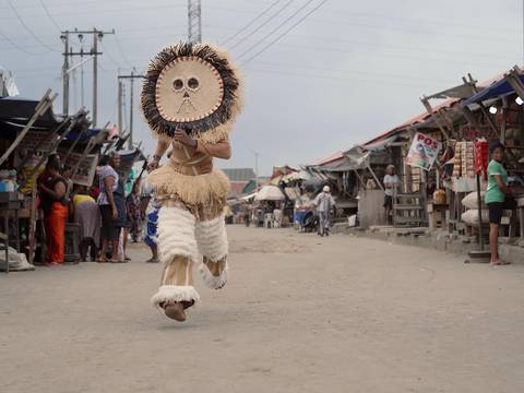  – Person in traditioneller Strohkostuem und Maskentanz auf einem Markt mit Staenden und Schaulustigen.