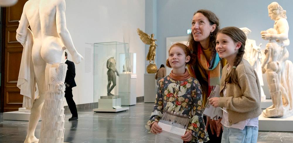 Ausstellungsbesuch im Alten Museum – Museumssaal mit Skulpturen. Drei Personen betrachten eine Statue. Hinten eine nackte männliche Figur in Marmor und ein Engel mit Flügeln aus Bronze.