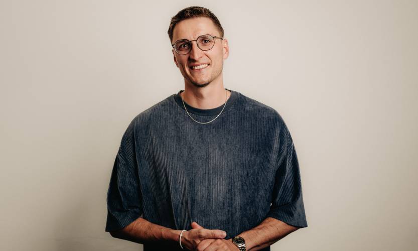 Johannes Steuding – Eine Person mit Brille und kurzem Haar trägt ein dunkelblaues Oberteil und eine Kette, lächelt vor hellem Hintergrund.
