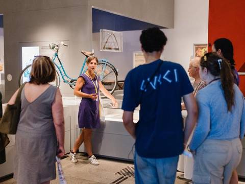  – Eine Guide ist in der Mitte des Bildes zu sehen und zeigt auf eine Glasvitrine. An den Rändern des Bildes sind Teilnehmende der Führung zu sehen. Im Hintergrund sieht man ein Fahrrad und eine Säge, die zwei Objekte der Ausstellung sind.