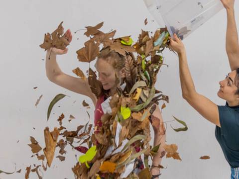 F(Olha)/Um(Blättern) – Zwei Personen, eine wirft Blätter und Papier aus einem Behälter auf die andere Person. Hintergrund neutral, beide scheinen aktiv zu sein.