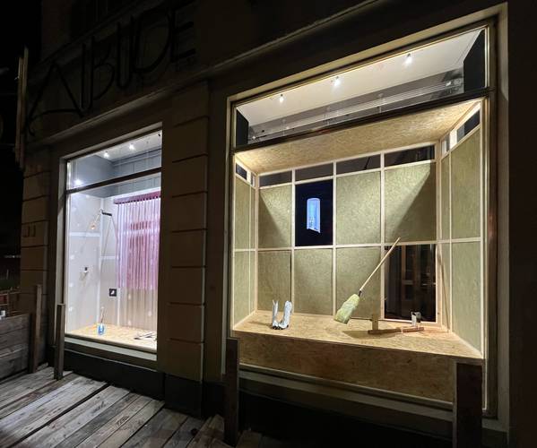Künstlerinnengespräch »A house and two windows« – Zwei Schaufenster bei Nacht, links mit rosa Lamellenvorhang, rechts Besen auf Holzboden. Schriftzug "Bude" über linkem Fenster.