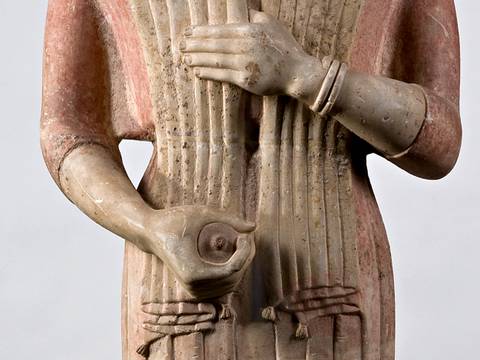  – Steinfigur mit roten und grauen Farbtönen, haelt ein Gefäß. Detaillierte Falten des Gewandes, Armbänder sichtbar. Helle Hintergrundfarbe.
