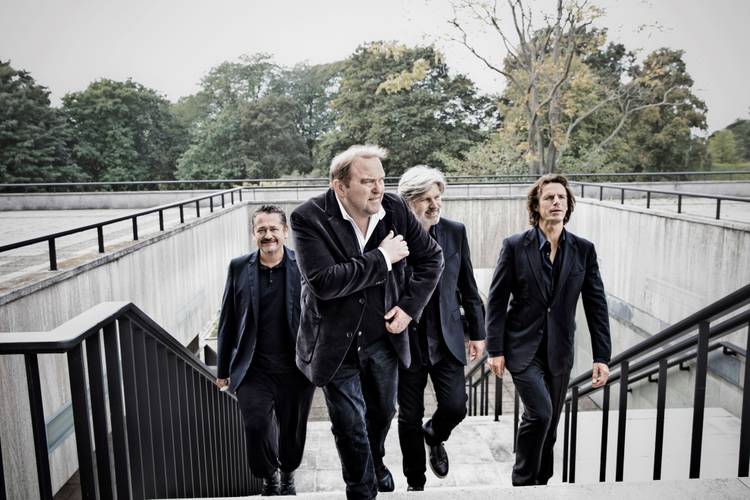 Gustav Peter Wöhler Band: LIVE ON STAGE – Vier Personen in dunklen Anzügen gehen im Freien eine Treppe hinauf. Im Hintergrund sind Bäume und ein bewölkter Himmel sichtbar.