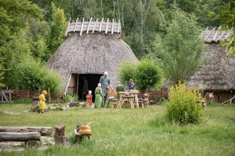 Die Darsteller:innen des Fördervereins Museumsdorf Düppel e.V. stellen mittelalterlichen Alltag nach. Besucher:innen können diesen live miterleben. – Ein Mann und mehrere Kinder in mittelalterlicher Kleidung halten sich vor einem rekonstruierten Haus auf dem Dorfplatz des Museumsdorf Düppel auf