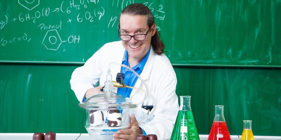 CheMagie Show – Person mit Brille und Laborkittel hinter einer Glasglocke. Hintergrund Tafel mit chemischen Formeln, darunter 6 H2O und Phenol.