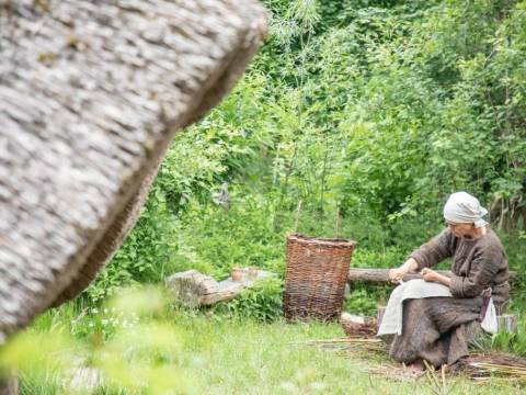 Eine Darstellerin des Fördervereins Museumsdorf Düppel e.V. bearbeitet Weidenruten, um daraus Körbe zu flechten. – Eine Frau sitzt auf einem Holzstumpf und bearbeitet mit einem kleinen Messer Weidenruten, daneben steht ein Weidenkorb
