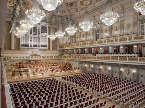  – Prunkvoller Konzertsaal mit großer Orgel, zahlreichen Kronleuchtern und leeren rot-weißen Stuhlreihen. Bühne mit leeren Musiker*innenplätzen.