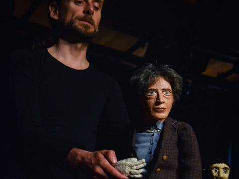 Wohl dem, der keine Heimat hat – Person hält Puppe mit grauen Haaren und braunem Sakko; Puppe sitzt auf Holzmöbel. Im Hintergrund zweite Puppe mit langem dunklem Haar.