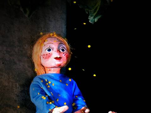  – Puppe in blauem Kleid sitzt mit ausgestreckten Beinen, verstreute gelbe Papierschnipsel, schwarzer Hintergrund.