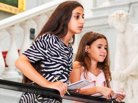 Veranstaltung in der Alten Nationalgalerie – Zwei Kinder auf Treppe in Museum, links Teenager in Zebrakleid, rechts Kind in rosa Top, beide halten Hefte, weiße Statue im Hintergrund.