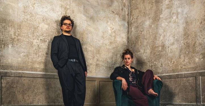 Eine Person steht in schwarzem Anzug, Hände in Taschen. Eine andere sitzt auf grünem Sessel in lässiger Kleidung. Hintergrund ist eine strukturierte Wand.