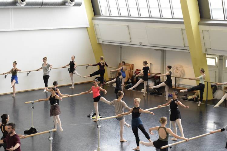 Mehrere Personen in Sportbekleidung trainieren an Ballettstangen in einem großen Raum mit Fenstern oben. Alle heben ein Bein an die Stange.